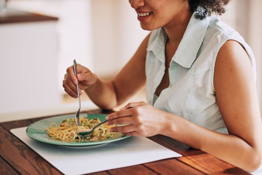 Une belle femme métisse mangeant des pâtes pour le dîner, assise à la table de la cuisine.