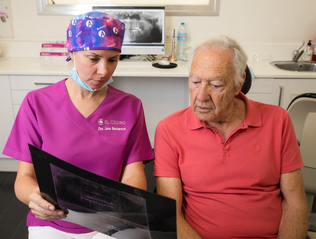 Une dentiste en blouse rose examine attentivement une radiographie dentaire avec un patient âgé assis à côté d'elle.